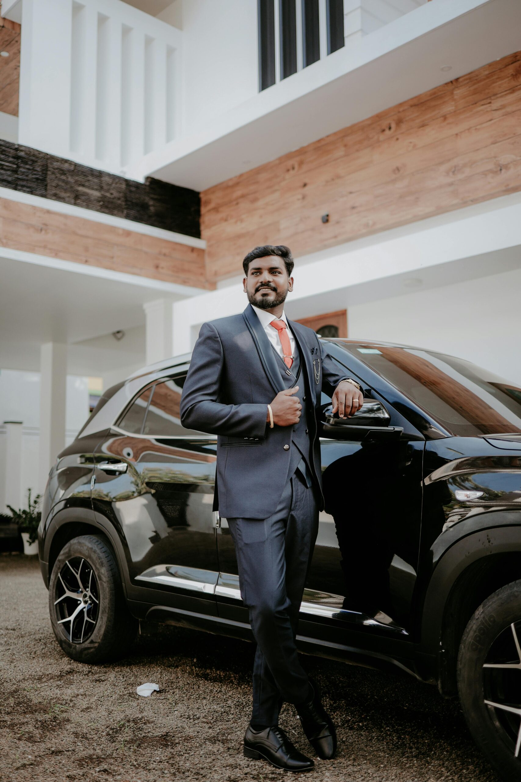 Stylish Indian groom in a blue suit confidently poses beside a sleek black car outdoors.